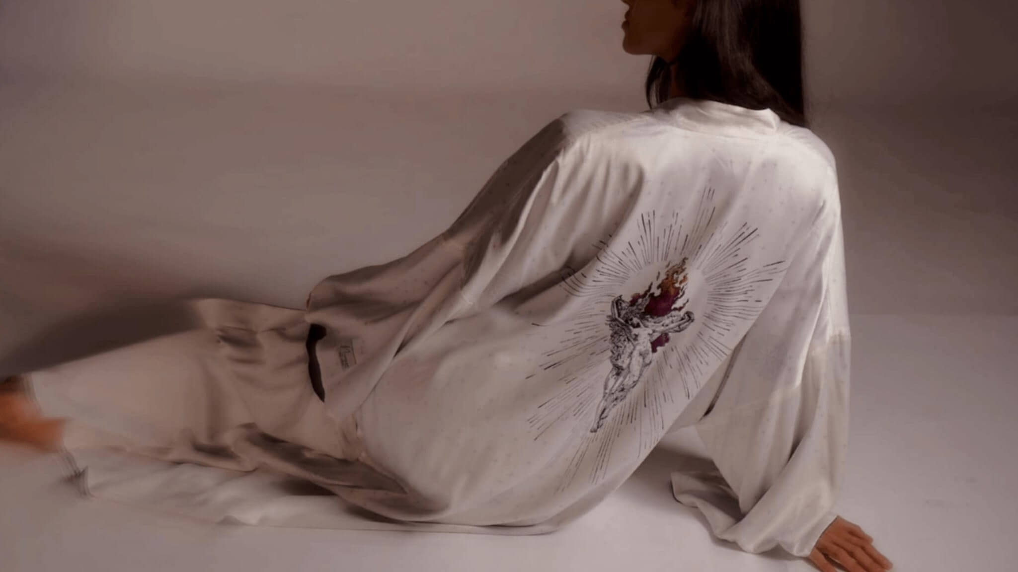 A woman lounging in the white silk Balcony Robe, featuring the exclusive art print on the back inspired by Symbolist painter Jean Delville.